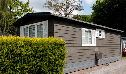 Ein kleines, einstöckiges graues Haus mit weißen Fensterläden, das teilweise von einem grünen Busch verdeckt wird, steht auf einem Kiesgrundstück am Mobilheimplatz Entenfangsee, mit Bäumen im Hintergrund.