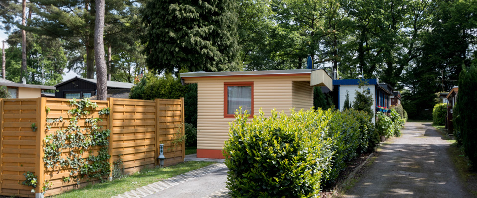 Ein kleines beigefarbenes Mobilheim mit roter Zierleiste steht inmitten von Grün, einem Holzzaun und Bäumen an einer schmalen gepflasterten Straße am friedlichen Mobilheimplatz Entenfangsee in einem ruhigen Wohnviertel.