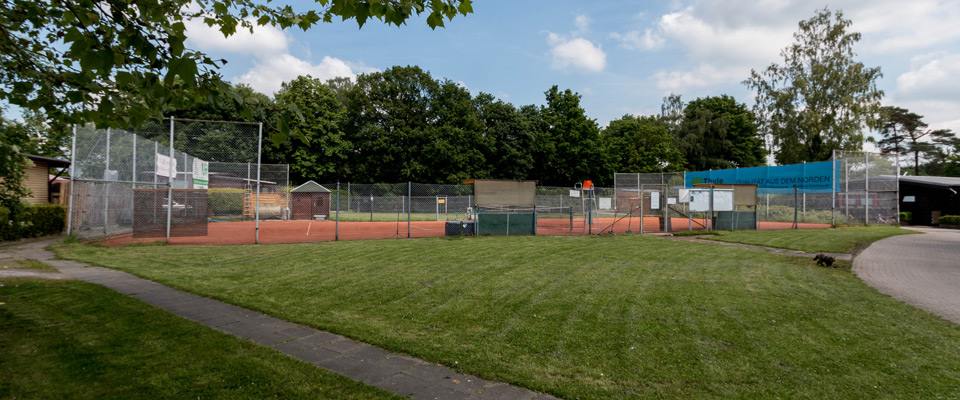 Die Tennisplätze mit rotem Sandbelag am Mobilheimplatz Entenfangsee sind von Zäunen und Bäumen umgeben, im Hintergrund sind bei teilweise bewölktem Himmel Schilder und ein kleines Gebäude zu sehen.