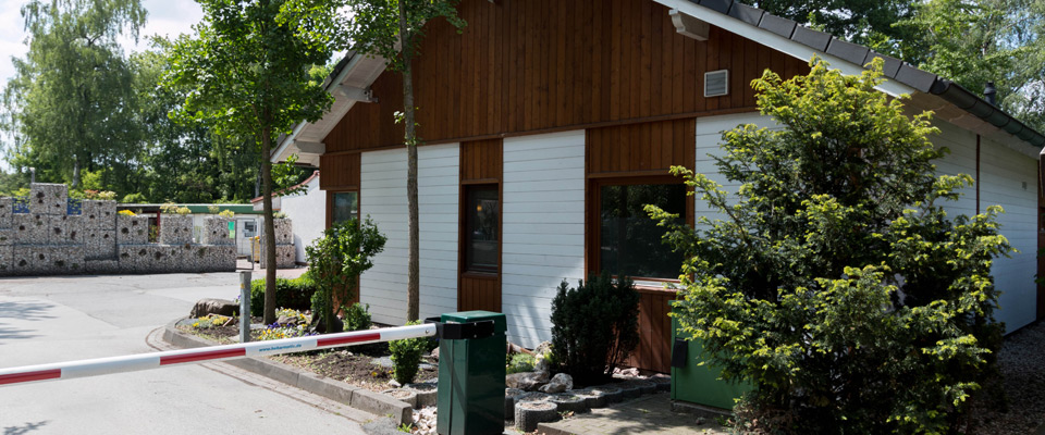 Hinter einer erhöhten Schranke am Mobilheimplatz Entenfangsee, umgeben von Bäumen und Sträuchern, steht an einem sonnigen Tag ein kleines Gebäude mit Holz und weißer Verkleidung.