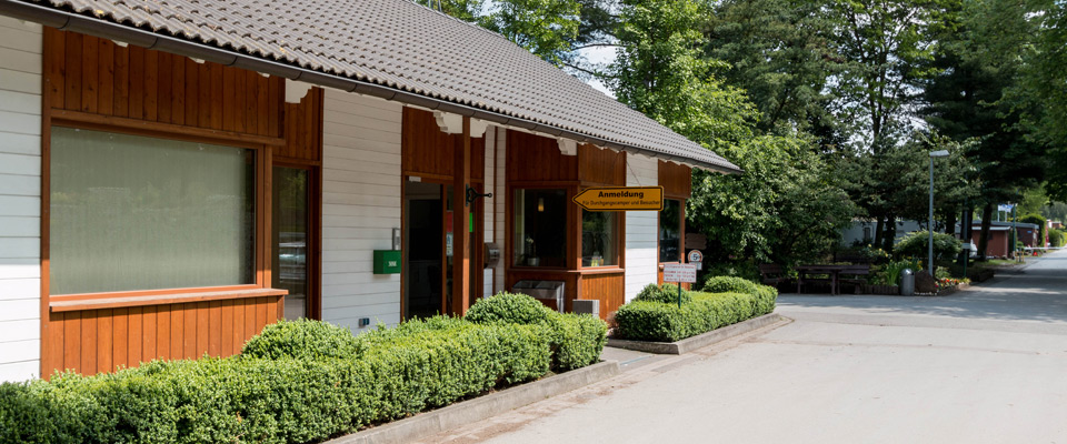 Ein einstöckiges Gebäude mit weißen Wänden und Holzverkleidung steht an einer gepflasterten Straße am Mobilheimplatz Entenfangsee, umgeben von grünen Sträuchern und Bäumen. Ein gelbes Schild hängt neben dem Eingang.