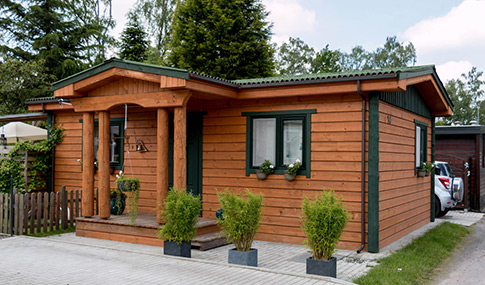 Charmantes kleines Holzhaus am Mobilheimplatz Entenfangsee mit begrüntem Dach, Veranda aus Holzstämmen, Topfpflanzen am Eingang und einem geparkten Auto, das teilweise rechts zu sehen ist.