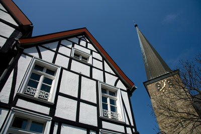 Ein traditionelles Fachwerkhaus steht neben einem hohen Kirchturm mit einer Uhr vor einem strahlend blauen Himmel.