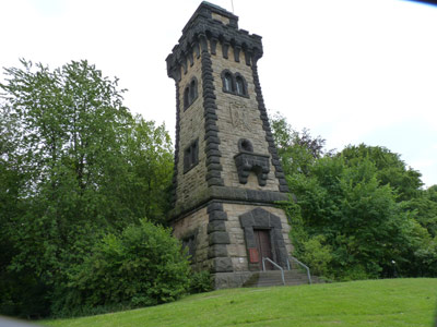 Aussichtsturm aus Stein mit Rundbogenfenstern und -tür, umgeben von Grün.