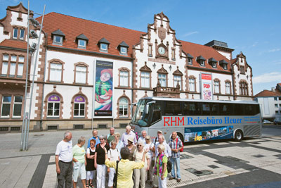 Eine Gruppe von Menschen steht vor einem Reisebus mit der Aufschrift RHM Reisebüro Heuer vor einem historischen Gebäude im europäischen Stil.