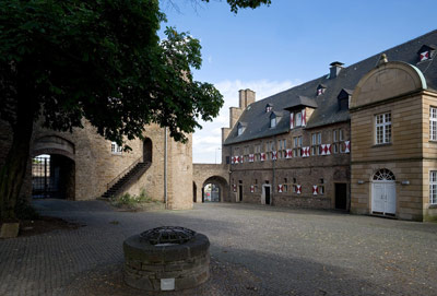 Ein steinerner Innenhof mit einem runden Brunnen, umgeben von historischen Gebäuden mit gewölbten Türen, verschlossenen Fenstern und Steinmauern unter einem teilweise bewölkten Himmel.