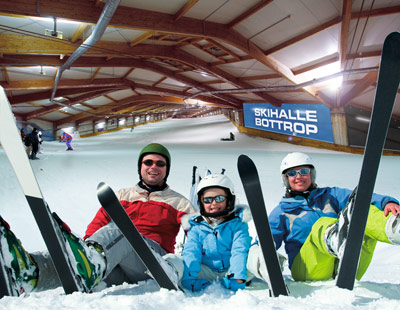 Drei Personen in Winterkleidung sitzen mit Skiern in einer Skihalle auf dem Schnee. Im Hintergrund ist das Schild der Skihalle Bottrop zu sehen.