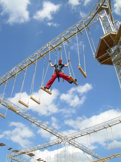Eine Person in Sicherheitsausrüstung navigiert in einem Hochseilgarten hoch über dem Boden unter einem blauen Himmel mit vereinzelten Wolken.