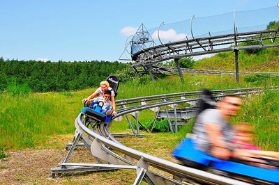 Auf einem grasbewachsenen Hügel unter blauem Himmel fahren Menschen auf einer alpinen Achterbahn aus Metall, wobei die unscharfe Bewegung die Geschwindigkeit anzeigt.