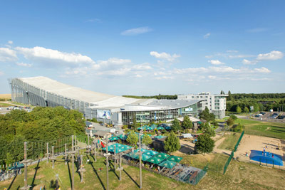 Ein großer überdachter Pistenkomplex mit einer Glasfassade, umgeben von Bäumen, Sitzgelegenheiten im Freien und einem Hochseilgarten unter blauem Himmel.