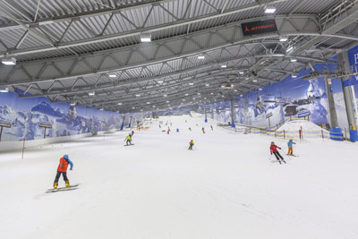 Skifahrer und Snowboarder in einer Skihalle mit Kunstschnee, unter einem hohen Metalldach mit Wandmalereien bedeckten Wänden.