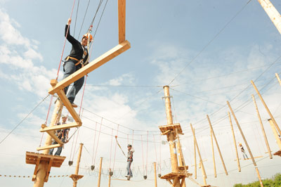 Unter freiem Himmel und bei blauem Himmel navigieren Menschen mit Klettergurten über erhöhte Seil- und Holzhindernisparcours.