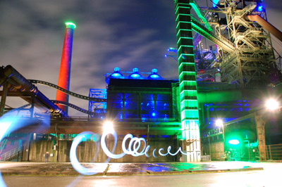 Ein nächtliches Industriegebäude wird mit grünen, blauen und roten Lichtern beleuchtet, mit wirbelnden Lichtspuren im Vordergrund.