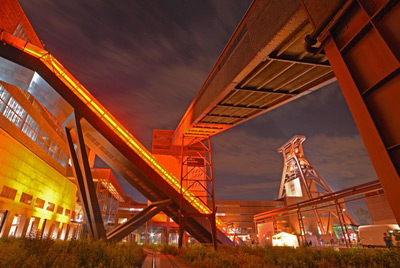 Industriekomplex bei Nacht mit beleuchteten Gehwegen und großen Stahlkonstruktionen unter einem bewölkten Himmel.